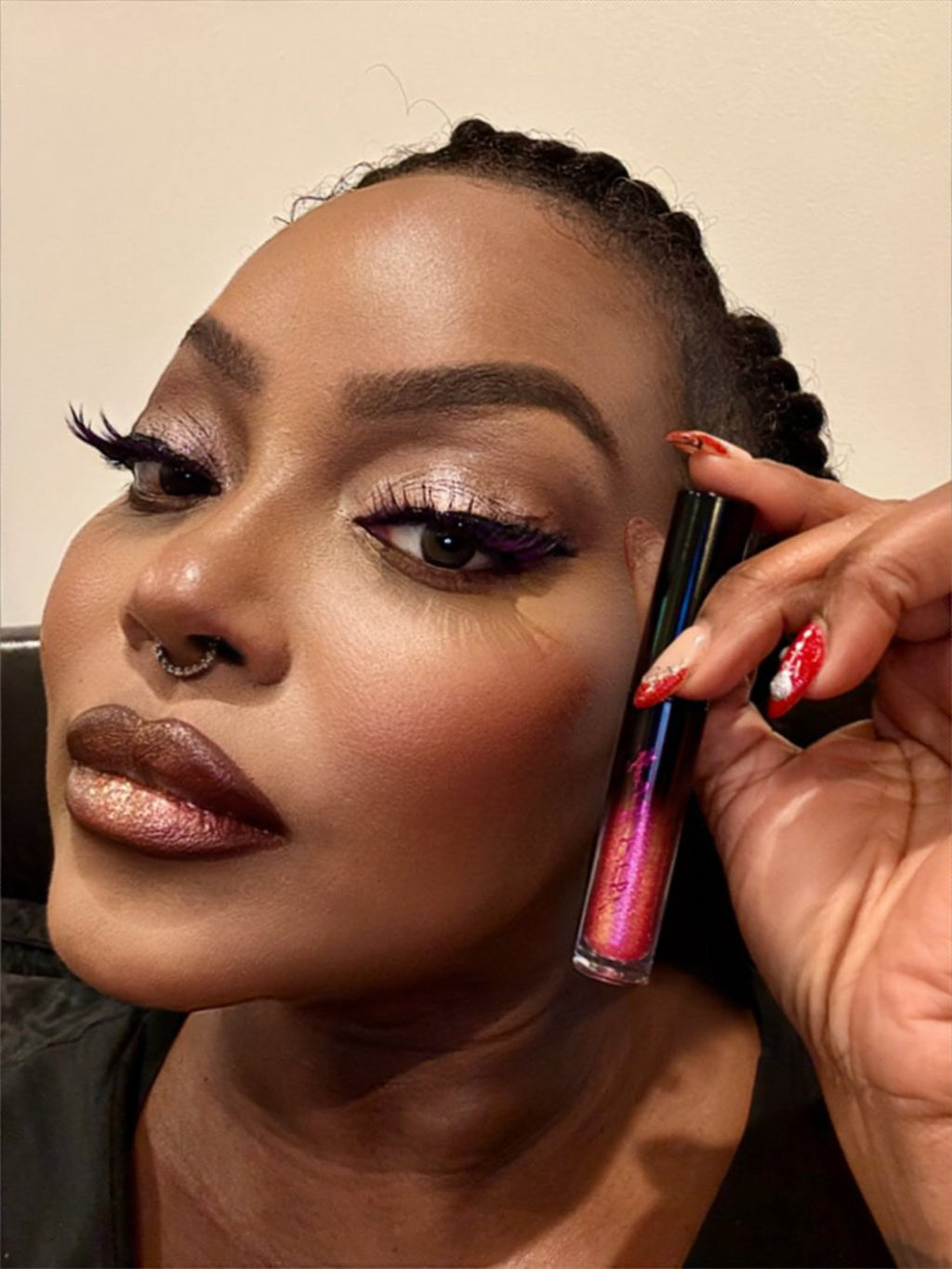 Woman holding red/black lip gloss, wearing purple-tinted false lashes, and having red and white decorated nails.